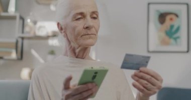 Akıllı telefon kullanan ve kredi kartı alışverişini evden yapan yaşlı bir kadın. Modern teknoloji ve tüketim kavramı.
