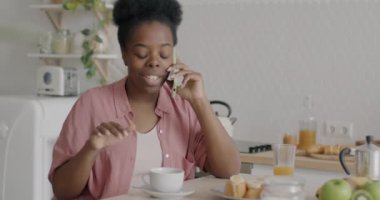 Çekici Afrikalı Amerikalı kadın cep telefonuyla konuşuyor kahkahalarla gülüyor mutfakta kahvaltıda kahvenin tadını çıkarıyor.