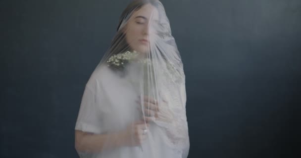 灰色の背景にプラスチックで包まれた花を嗅ぐ敏感な若い女性のスローモーション 女性性と性格の概念 — ストック動画