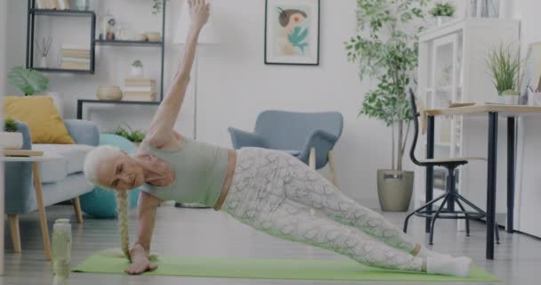 穿着运动服的退休女运动员在家里做瑜伽 在垫子上享受天平的肖像 精力充沛的老年妇女独自接受培训 — 图库视频影像