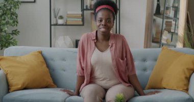 Evdeki kanepede oturmuş kameraya gülümseyen güzel Afro-Amerikan kadının yavaş çekimde portresi. Halk ve yerli geçmiş kavramı.