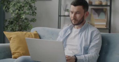 Evde rahat bir kanepede oturan dizüstü bilgisayarla çalışan melez bir adamın portresi. Modern teknoloji ve serbest çalışma kavramı.