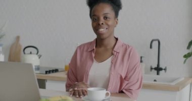 Mutfak masasında dizüstü bilgisayarla oturmuş kameraya gülümseyen Afro-Amerikan kadının yavaş çekim portresi. Ev ve elektronik aygıt kavramından çalışma.