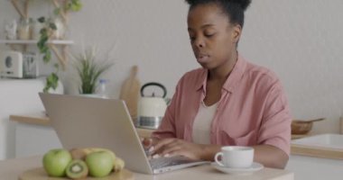 Hırslı Afro-Amerikalı kadın evde mutfaktaki masada bilgisayarla yazı yazıyor ve iş aktivitelerine odaklanmış durumda. İnsanlar ve uzak iş konsepti.