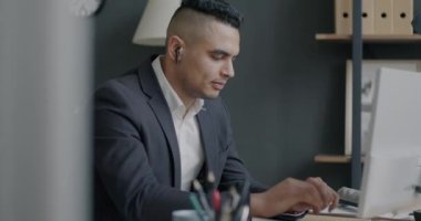 Neşeli Orta Doğulu iş adamı kablosuz kulaklık takıyor müzik dinliyor ve ofiste bilgisayarla çalışıyor. İnsanlar ve işyeri konsepti.