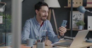 Neşeli genç adam konuşuyor ve ofisteki akıllı telefonuyla el sallıyor. İletişim ve işletme sahibi kavramı.