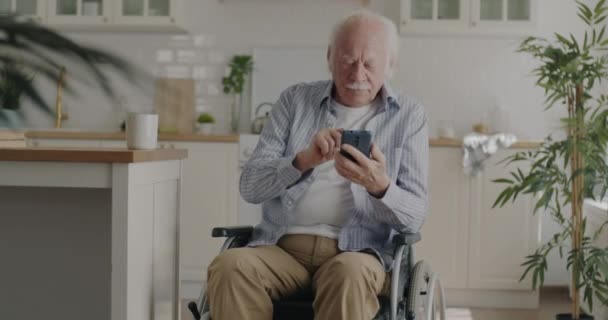 无忧无虑的老人用智能手机享受着社交媒体坐在厨房的轮椅上 现代技术与退休人员概念 — 图库视频影像