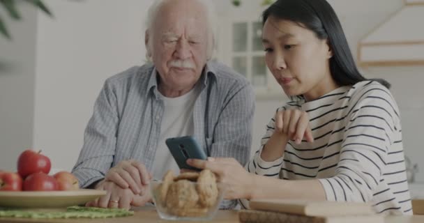 Neşeli Hemşire Yaşlı Adama Akıllı Telefon Ekranını Gösteriyor Evde Mutfakta — Stok video