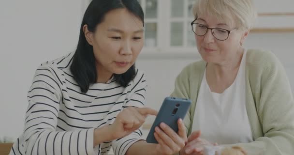 退休女士在家里的厨房里学习使用智能手机应用程序与年轻女性志愿者交谈 现代技术和老年人援助概念 — 图库视频影像