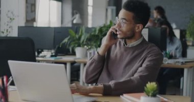 Ofis çalışanı Afrikalı Amerikalı cep telefonuyla konuşuyor ve dizüstü bilgisayarla çalışıyor. İş görüşmesi ve elektronik aygıtlar kavramı.