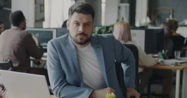 Açık uzay ofisindeki kameraya bakan kendine güvenen bir adamın yavaş çekim portresi. Zarif takım elbiseli bir işçi başarılı bir kariyerin tadını çıkarıyor..