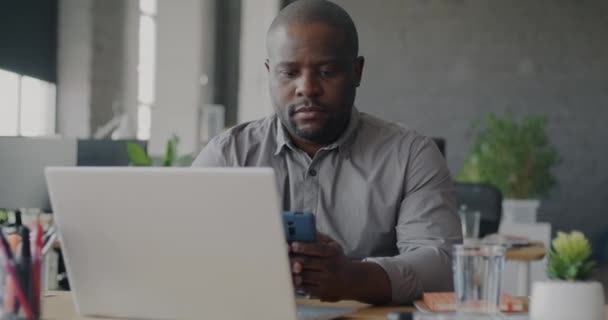職場での企業コミュニケーションで忙しいスマートフォンやノートパソコンを使用して深刻なアフリカ系アメリカ人のオフィスワーカー 現代のテクノロジーとビジネス界の概念 — ストック動画