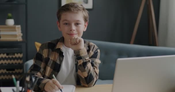 一个聪明的孩子坐在桌旁 手里拿着笔记本和笔 微笑着看着家里的相机 教育和儿童概念 — 图库视频影像