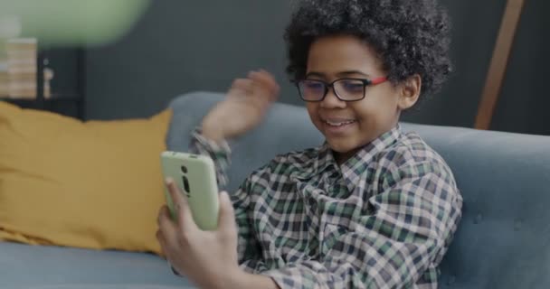 可愛い男の子がオンラインビデオ通話をしたり アパートでスマートフォンを使って手を振っています 通信とデバイスの概念 — ストック動画