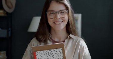 Evde elinde çizim defterleri tutarak gülümseyen neşeli serbest kız illüstratörün yavaş çekimleri. Pozitif duygular ve uzak iş kavramı.