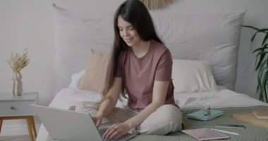 Dizüstü bilgisayarla çalışan serbest çalışan genç bir kadın evde yatakta oturuyor. Modern teknoloji ve apartman konseptinde uzaktan çalışma.