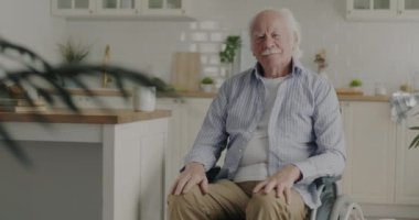 Evde, modern tekerlekli sandalyede oturan ve düşünceli yüzüyle kameraya bakan sessiz, yaşlı bir adamın ağır çekimde portresi. Engelliler ve yaşam tarzı konsepti.