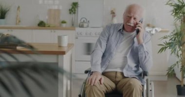Neşeli yaşlı adam cep telefonuyla konuşuyor ve gülüyor. Evdeki mutfakta tekerlekli sandalyede oturuyor. İletişim ve emeklilik kavramı.