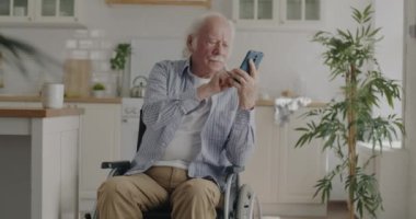Neşeli emekli adam akıllı telefon ekranına dokunuyor ve tekerlekli sandalyede otururken gülümsüyor. Gadget ve mutlu emeklilik kavramı.