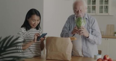 Genç bayan konuşurken ve evde akıllı telefon kullanırken, yaşlı adam kağıt torbadaki sağlıklı organik yiyecekleri boşaltıyor. Ürün teslimatı ve beslenme konsepti.