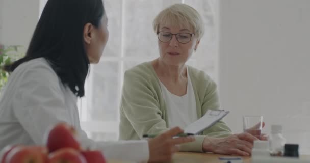 退休妇女在公寓里与穿着白衣的女医生讨论健康问题 在家中医疗记录中谈话和写作的妇女 — 图库视频影像