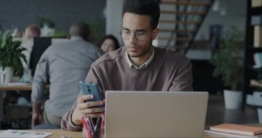 Hırslı Afro-Amerikalı adam dizüstü bilgisayar kullanıyor ve akıllı telefonla mesaj yazıyor. Ofisteki müşterilerden emir alıyor. İnsanlar ve kurumsal iletişim konsepti.