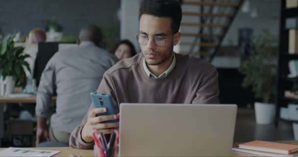 野心的なアフリカ系アメリカ人の男性は オフィスでクライアントからの注文を受けてスマートフォンでノートパソコンやテキストメッセージを使用しています 人と企業のコミュニケーションの概念 — ストック動画