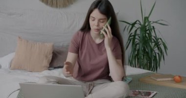Genç bayan cep telefonuyla konuşuyor ve bilgisayarla çalışıyor. Uzaktan kumandalı işten zevk alıyor. Evde yatakta oturuyor. İnsanlar ve serbest çalışma kavramı.
