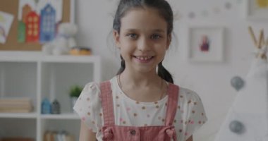 Evde el sallayarak gülümseyen neşeli küçük kızın yavaş çekimde portresi. Mutlu çocukluk ve pozitif duygu kavramı.