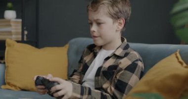 Oyun oynayan neşeli bir çocuk evde koltukta oturup zaferin tadını çıkarırken joystickle gülüyor. Modern teknoloji ve çocuk konsepti.