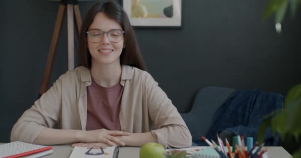 屋内の自宅でカメラを見て笑顔イラストでテーブルに座っている美しい若い女性アーティストのスローモーションの肖像画 人と趣味の概念 — ストック動画