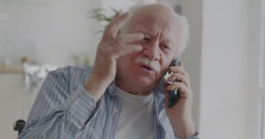 Duygusal yaşlı bir adam cep telefonuyla konuşuyor ve evde el kol hareketi yapıyor. Kablosuz iletişim teknolojisi ve emeklilik kavramı.