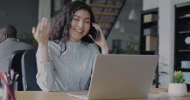 Neşeli genç bayan yönetici cep telefonuyla konuşuyor ve iş yerinde dizüstü bilgisayarla çalışıyor. İş iletişimi ve teknoloji kavramı.