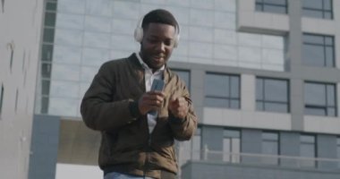 Kaygısız Afrikalı Amerikalı öğrenci portresi kablosuz kulaklıklar ve akıllı telefonlardan müzik dinliyor açık havada eğleniyor..