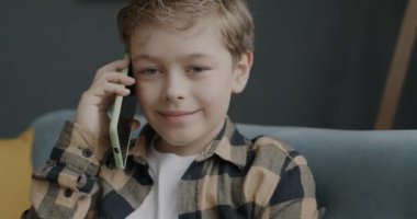 Modern apartmandaki koltukta oturan neşeli çocuk cep telefonuyla konuşuyor ve gülümsüyor. Eğlenceli sohbetler ve mutlu çocukluk kavramları.