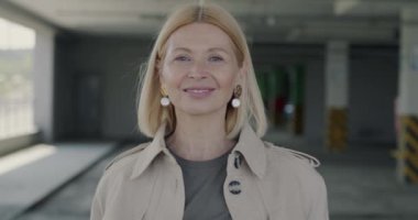 Otoparkta tek başına durup kameraya bakarken gülümseyen neşeli olgun bir kadının yavaş çekim portresi. Halk ve şehir yaşam tarzı kavramı.