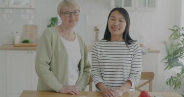 Neşeli insanların portresi genç Asyalı kadın ve kıdemli beyaz kadın mutfakta ayakta gülümsüyor. Pozitif duygu ve yaşam tarzı kavramı.