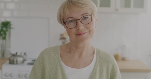 台所の自宅でカメラを見て笑顔メガネを身に着けている陽気な先輩女性のスローモーションポートレート 老人と家の所有者の概念 — ストック動画