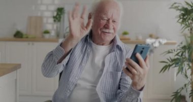 İnternette video görüşmesi yapan mutlu kıdemli bir adam mutfaktaki evde el sallıyor. İletişim ve emeklilik kavramı.