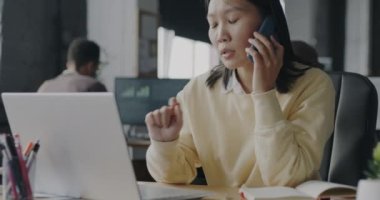 Genç Asyalı bayan ofis çalışanı cep telefonuyla konuşuyor ve bilgisayarla içeride çalışıyor. Arka planda iş faaliyetlerine katılan meslektaşlar.