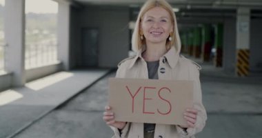 Evet tabelasını tutan neşeli bir kadının portresi, şehirdeki otoparkta duran gülümseyen bir anlaşmayı ifade ediyor. Pozitif duygu ve kentsel yaşam tarzı kavramı.