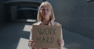 Elinde ilham verici bir tabela tutan olgun bir kadının ağır çekimde çekilmiş portresi. Kentsel arka plana karşı çok çalış. Kariyer ve motivasyon kavramı.