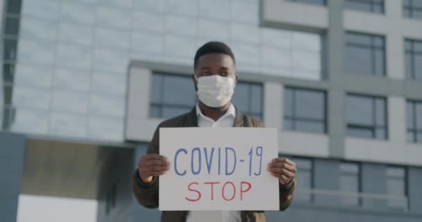 ストップCovid 19バナーを保持街の通りを歩く医療面マスクを身に着けているアフリカ系アメリカ人男性のスローモーション肖像画 人々とパンデミックの概念 — ストック動画