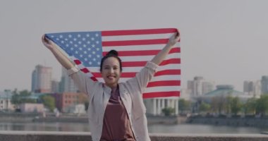 Modern şehir caddesinde elinde Amerikan bayrağı sallayan mutlu bir kadının yavaş çekimde portresi. Amerika Birleşik Devletleri ve insanlar konsepti.