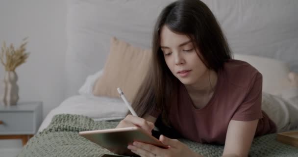 タブレットを描き アパートのベッドでリラックスした笑顔で働く幸せな女子学生 現代のテクノロジーとミレニアルの概念 — ストック動画