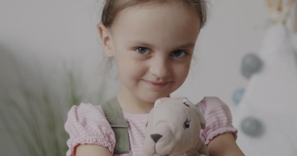 アパート内のカメラを屋内で見て笑顔柔らかいおもちゃのウサギを保持甘い女の子のスローモーションポートレート 人々と幸せな子供時代のコンセプト — ストック動画