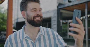Akıllı telefondan konuşan neşeli bir adam çevrimiçi video görüşmesi yapıyor açık havada konuşuyor ve gülüyor. Gençler ve iletişim konsepti.