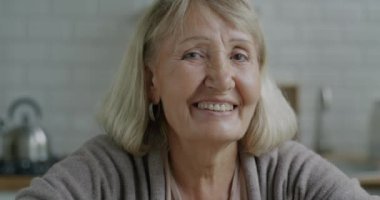 Dairenin mutfağında kameraya bakarak gülümseyen sarı saçlı güzel yaşlı bir kadının yakın plan portresi. Emeklilik ve mutlu yaşlılar konsepti.