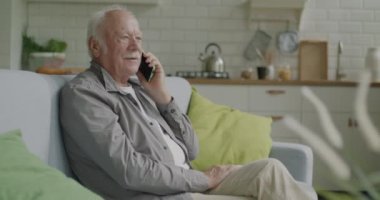 Yaşlı adam cep telefonuyla konuşuyor. Sıcak bir dairede koltukta otururken şaşkınlıkla gülümsüyor. İletişim ve emekliler konsepti.
