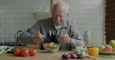 Yaşlı bir adamın sebzeleri kaseye karıştırıp salata pişirmesi ve evde yemek yerken gülümsemesi. Beslenme ve yaşlılar konsepti.